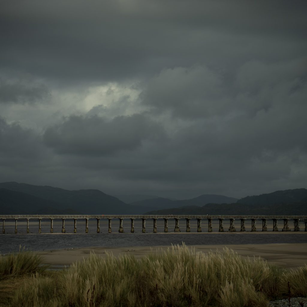 Meghan Worden - A bridge across a river at low tide with cloudy skies above hillside and grasses in the foreground