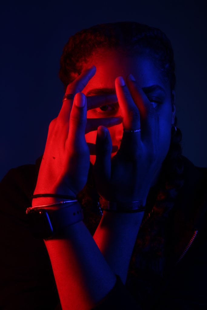 Caitlin Fryer - Portrait of a person in red and blue light against a black background, with hands in front of their face