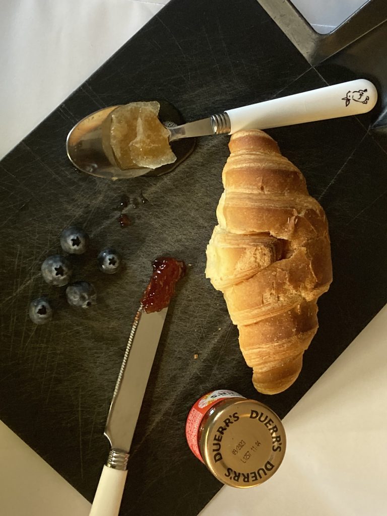 Gracie Eddy - A croissant on a chopping board with blueberries, jam, a knife and spoon