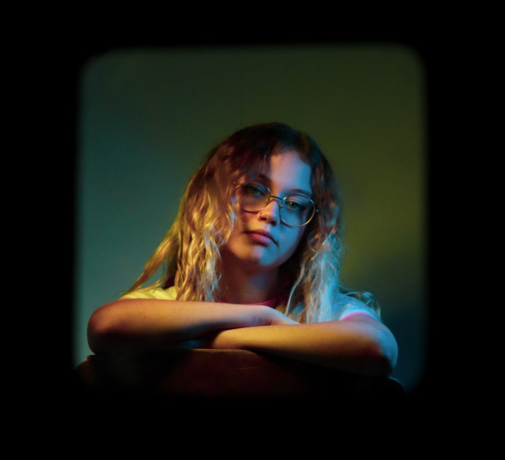 Holly Perkowski - A girl with long curly hair and glasses sits with her arms crossed, framed by a green square against a black background