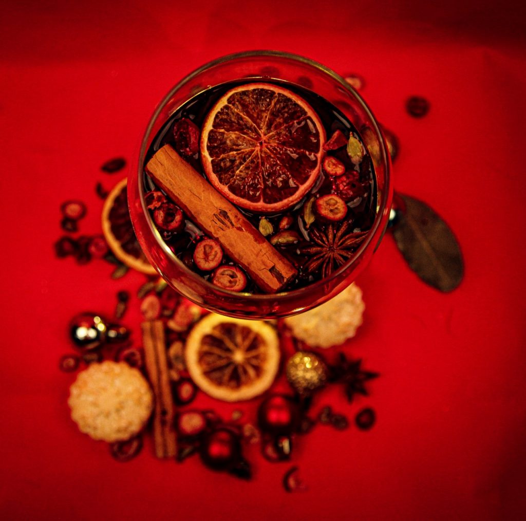 Holly Perkowski - A glass of mulled wine with fruit and spices in the liquid and surrounding the glass on a red background