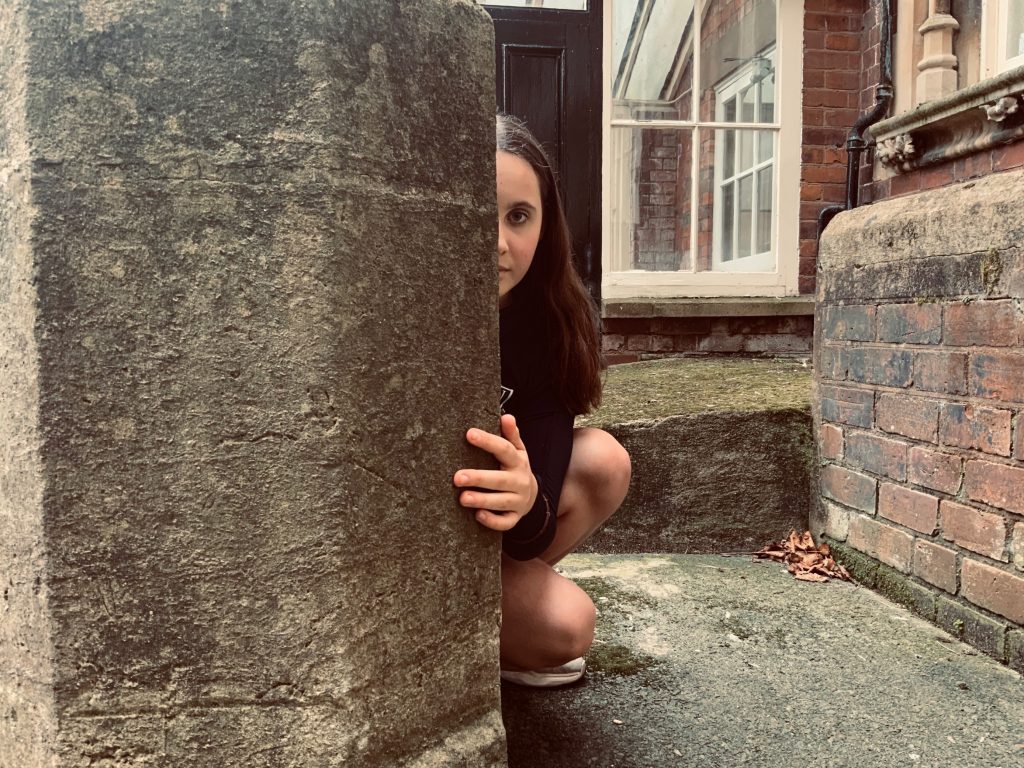 Imogen Hobbs - A young girl crouched down hiding behind a stone pillar in front of a building