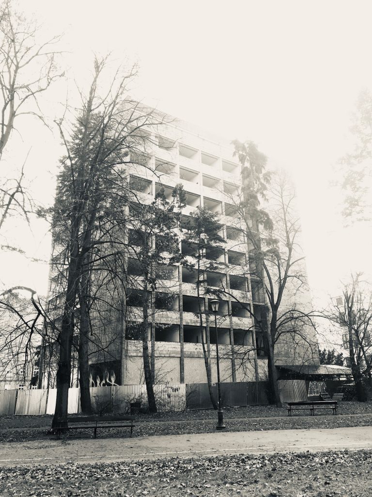 Iwo Kozicki - Monochrome image of an abandoned modern apartment building taken from a park with trees partially covering the building
