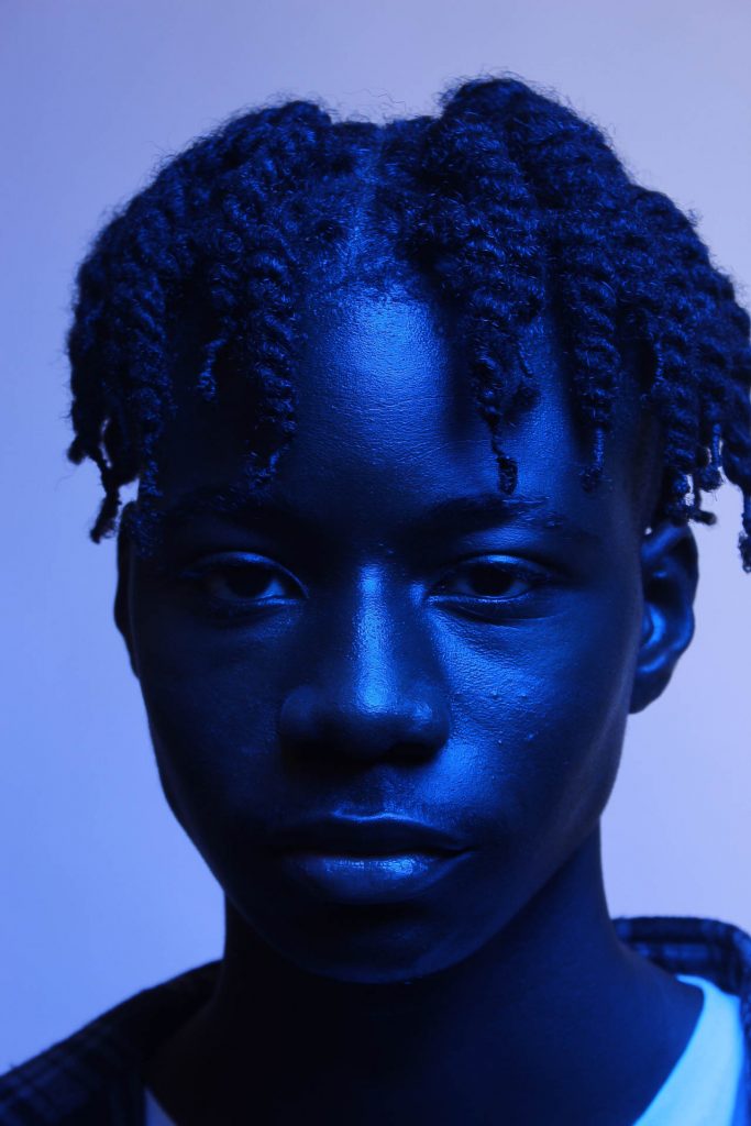 Leilani Drummey - A boy with dark skin and hair is lit by blue light on a pale background