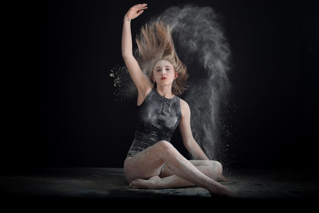 Lydia Jones - A girl sits throwing white powder in the air against a black backdrop with her arm raised and dust falling around her