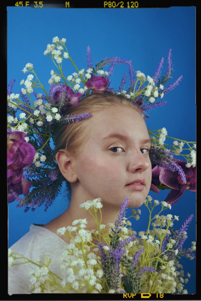 Maria Ignatieva - A young girl with a crown and bouquet of purple and white flowers photographed against a blue backdrop