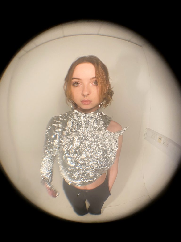 Mia Yearn - A girl wearing a tin foil top framed in a fish-eye lens with a black background surrounding the white circle