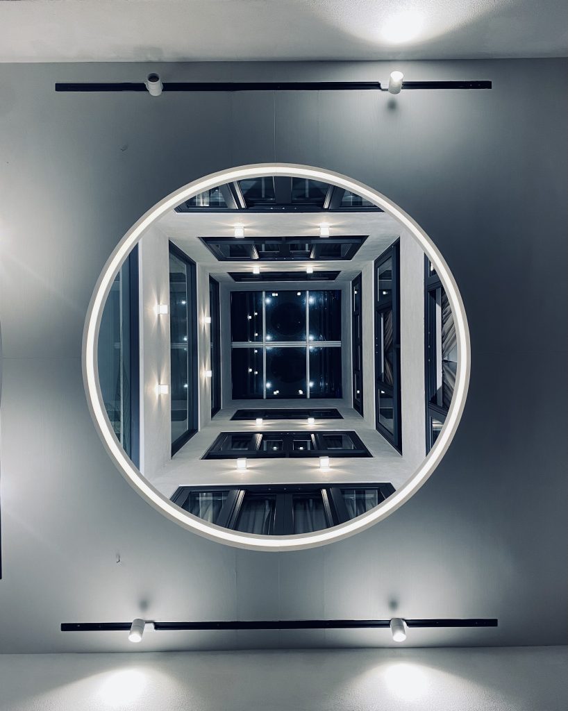Nick Chan - View through a circular hole in a ceiling looking up through multiple floors of a building with lights and windows