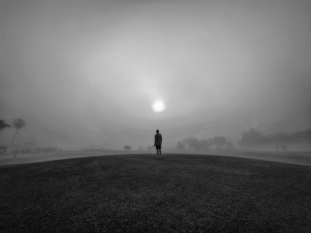 Rohit Jayade - Monochrome image of a man stood on a mound of grass with blurred skyline showing trees and the sun