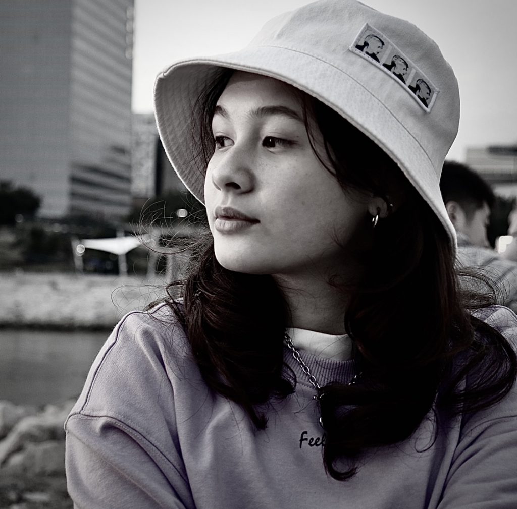 Salomé De Sousa - Monochrome image of a young girl wearing a white bucket hat with dark hair against a city backdrop