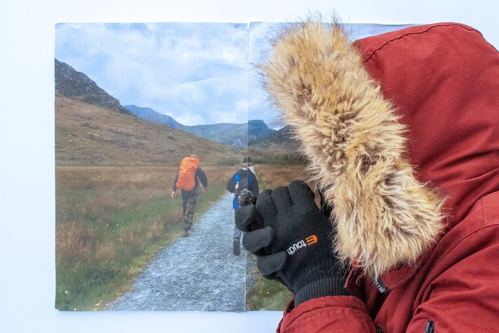 Thomas Chiplin - Person wearing a red parka coat with fur hood and black gloves stood by a printed picture of two hikers walking along a mountain path