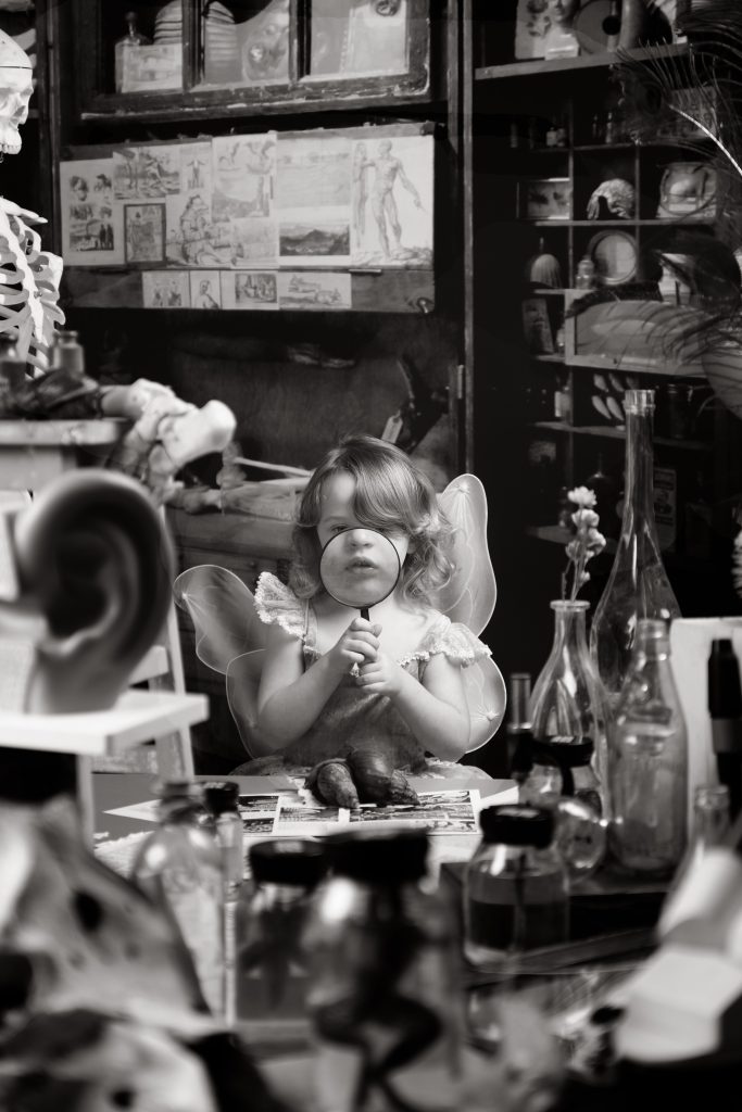 Zoe Kingston - Monochrome image of a toddler in fairy fancy dress holding a magnifying glass to her face, stood in a room filled with unusual objects