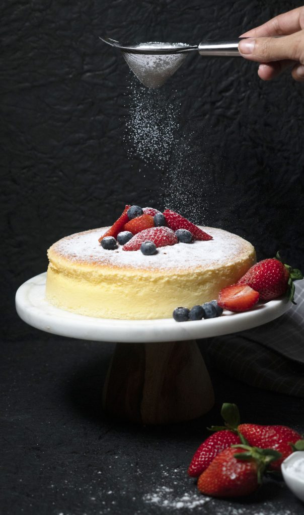 Aamna Junaid - Sponge cake dusted with sugar and topped with berries against a black background.
