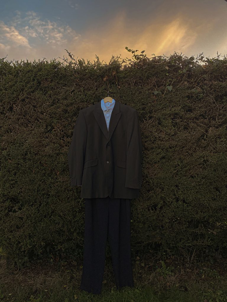 Aliyah Jay - A suit hanging on a tall dense hedge at dusk.