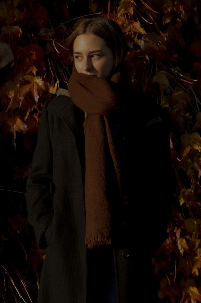 Aliyah Jay - A girl wearing a thick rust coloured scarf against a backdrop of leaves at dusk.
