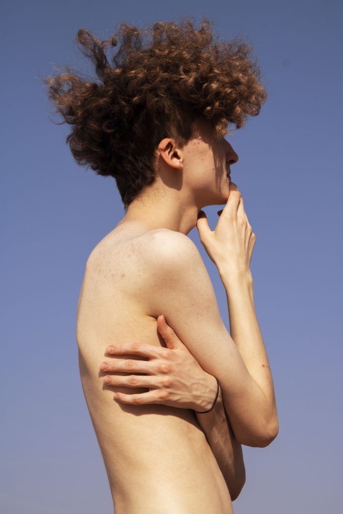 Amy Boyd - A bare-skinned young person with afro hair stares into the distance against blue sky.