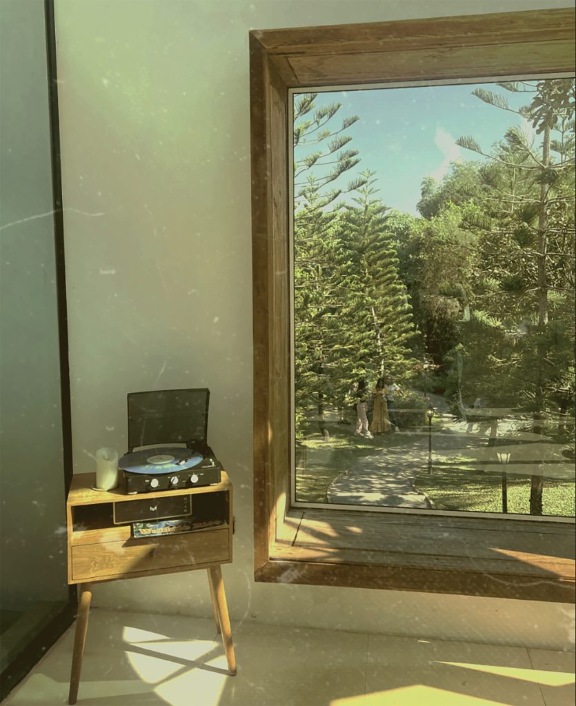 Astrid Htet - Sepia toned view of woodland from a large wooden window, with a record player on a side table beside it.