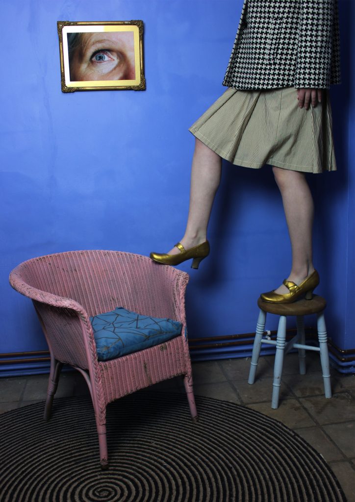 Bunny Edwards - Legs of a person in a grey skirt and gold heels standing across a stool and rattan chair against a deep blue wall.