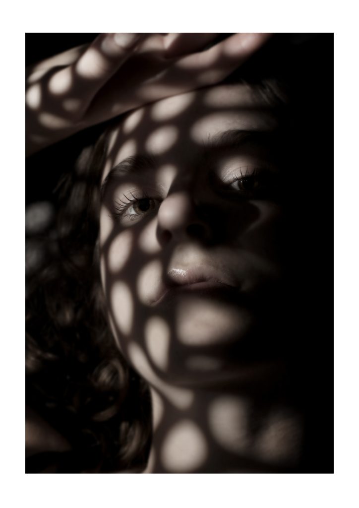 Ella Martin - Portrait of a young woman lit by dappled white light against a black backdrop.