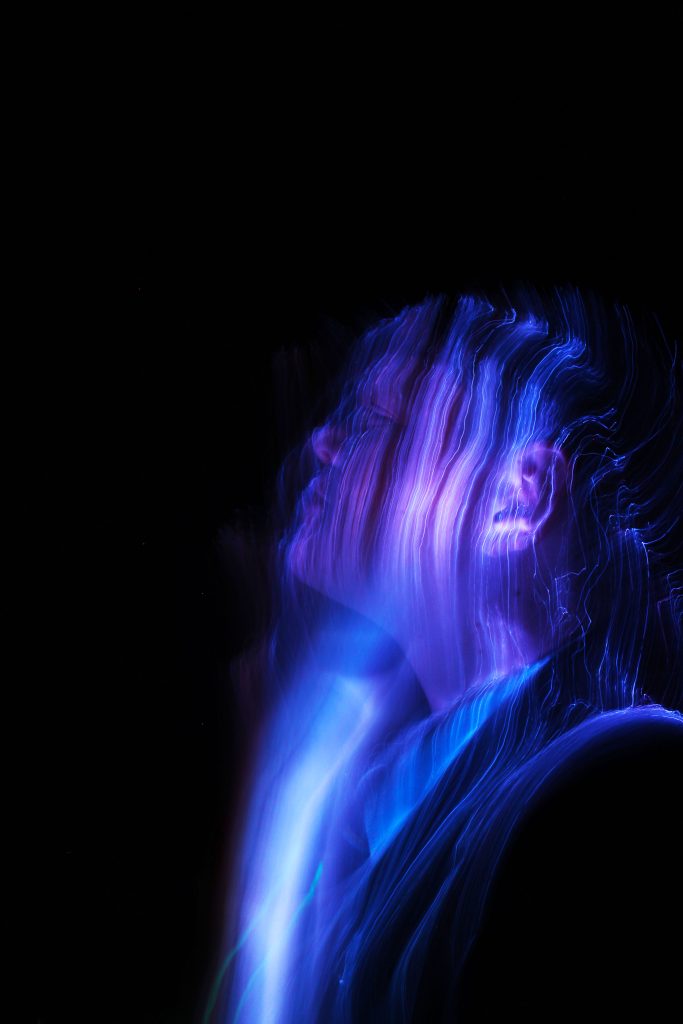 Laura Duffy - A person with long hair lit by streaks of neon purple light against a black backdrop.