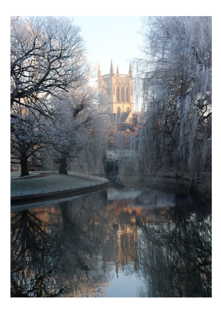 Sam Clarke - View of a cathedral from the banks of a river through frost covered leafless trees.