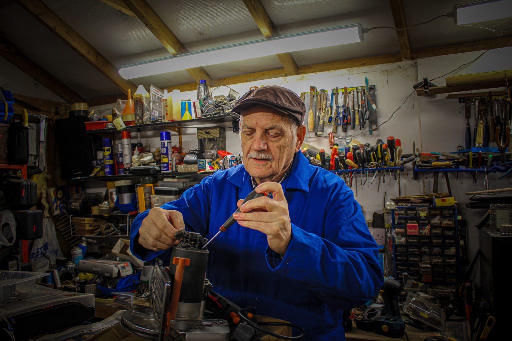 Scarlett Turner - An older man in a blue boiler suit and camp works on a project in his garage, surrounded by tools.