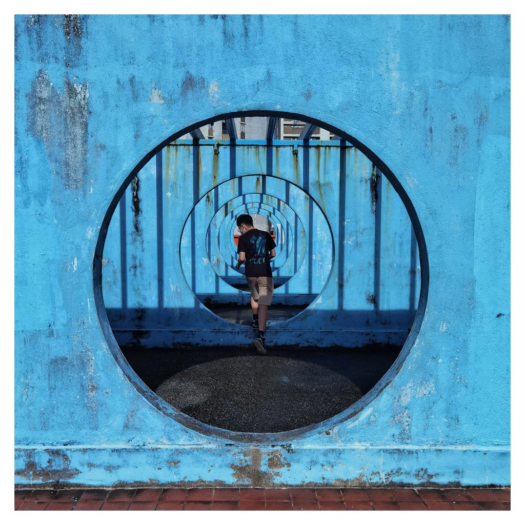 Xu Ka-ngai - Image of a person walking through a row of circular openings in blue walls going off into the distance.
